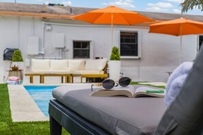Eine Pool-Lounge mit einer Couch, einem offenen Buch und Sonnenbrillen, sowie zwei orangefarbenen Sonnenschirmen.