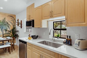 A kitchen with wooden cabinets, a microwave, a sink, a window, and a dining area with chairs and a table. - Casa Palma | Pool | Outdoor Fun | Near Beaches (Fort Lauderdale)