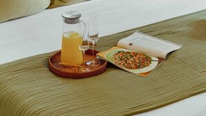 Ein ordentlich gemachtes Bett mit Kissen, ein Glas Orangensaft, ein Teller Essen und ein Buch auf einem Nachttisch.