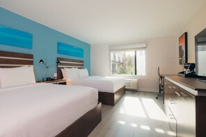 Ein Hotelzimmer mit zwei Betten, einem Schreibtisch, einem Stuhl und einem Fenster mit Blick auf Gebäude.