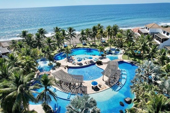 A resort with a central pool, palm trees, and a view of the ocean.