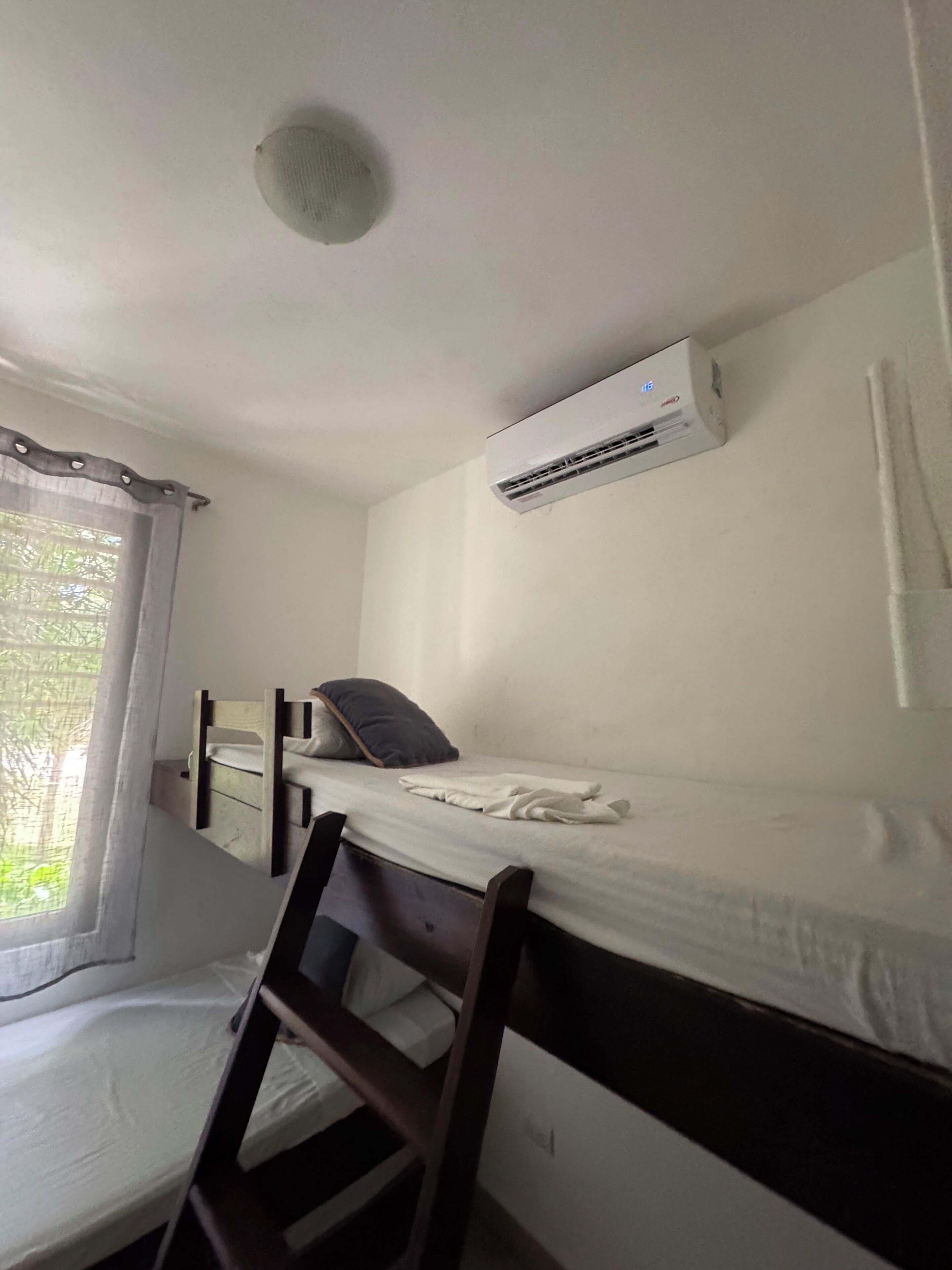 Una cama individual con estructura de madera, colchón blanco y almohada. Un equipo de aire acondicionado está montado en la pared sobre la cama.