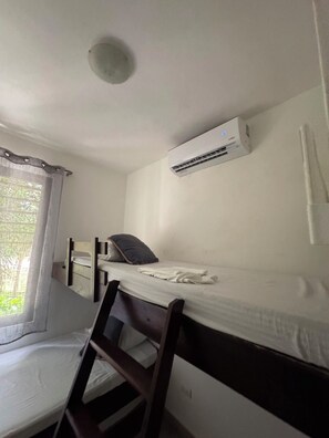 A single bed with a wooden frame, a white mattress, and a pillow. An air conditioning unit is mounted on the wall above the bed.