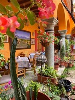 Un espacio exterior para comer con mesas y sillas, rodeado de plantas en macetas y un pilar de piedra.