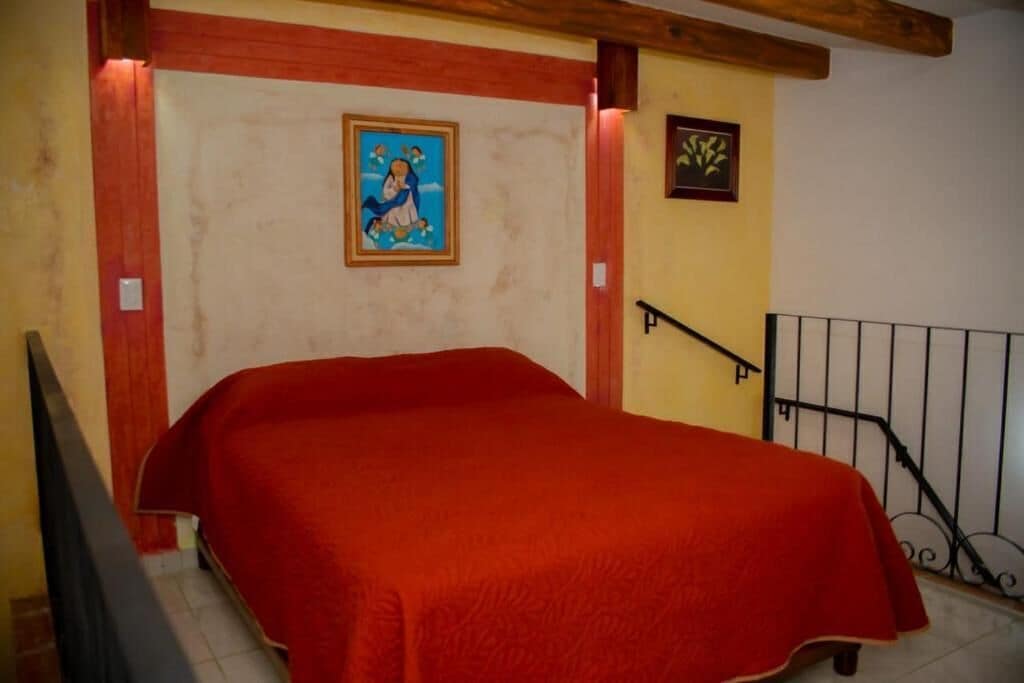 A room with a bed, a wooden ceiling, a wall with a framed picture, and a staircase with a railing.