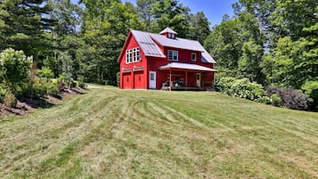 Una casa roja con un porche delantero amplio y techo inclinado, rodeada de un césped cuidado y árboles frondosos.