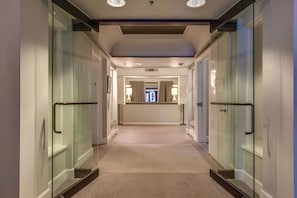 Corredor moderno de hotel com portas de vidro, luminária central e vista para um quarto com janela.