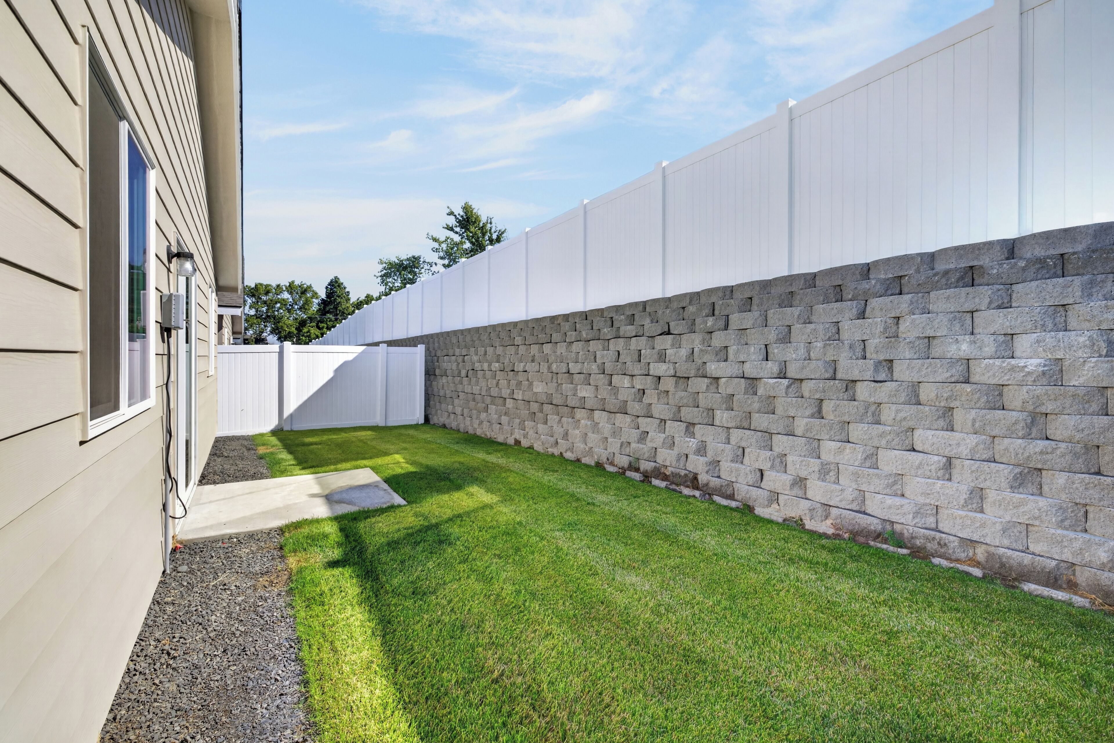 A backyard with a paved area, a stone wall, and a white fence.