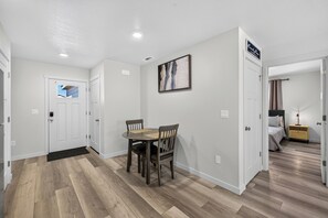 Quarto de hotel com piso de madeira, uma mesa de jantar com cadeiras, uma cama e uma mesa de cabeceira com abajur.