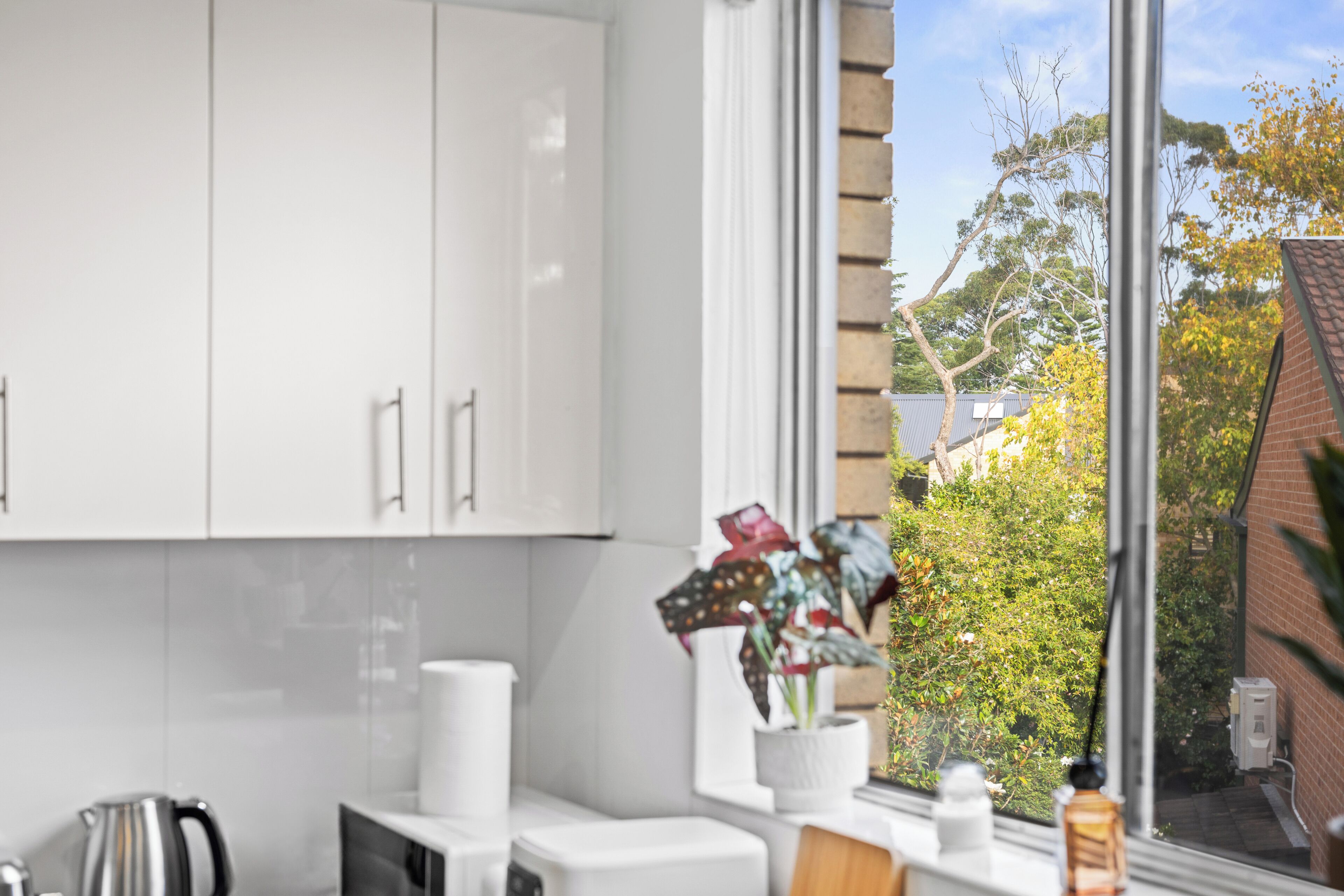 Une cuisine moderne avec des meubles blancs, une bouilloire et une fenêtre donnant sur des arbres et un immeuble.