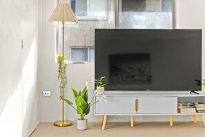 Un saló modern amb un televisor de pantalla plana sobre un moble blanc, una làmpada de peu i una planta en test.