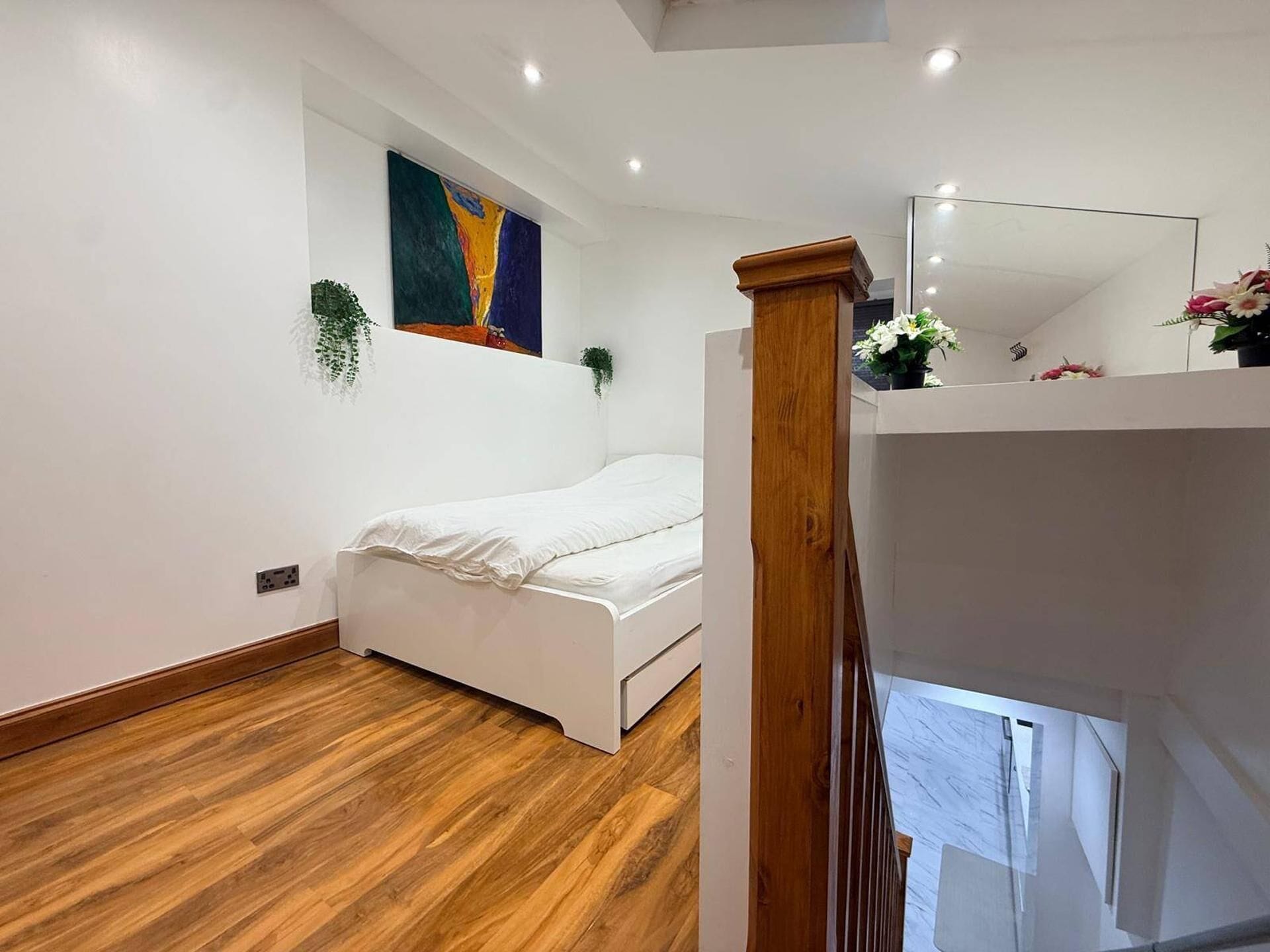 Un dormitorio con piso de madera, una cama blanca, un cuadro en la pared y una planta pequeña.