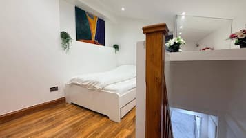 A bedroom with a wooden floor, a white bed, a painting on the wall, and a small plant.