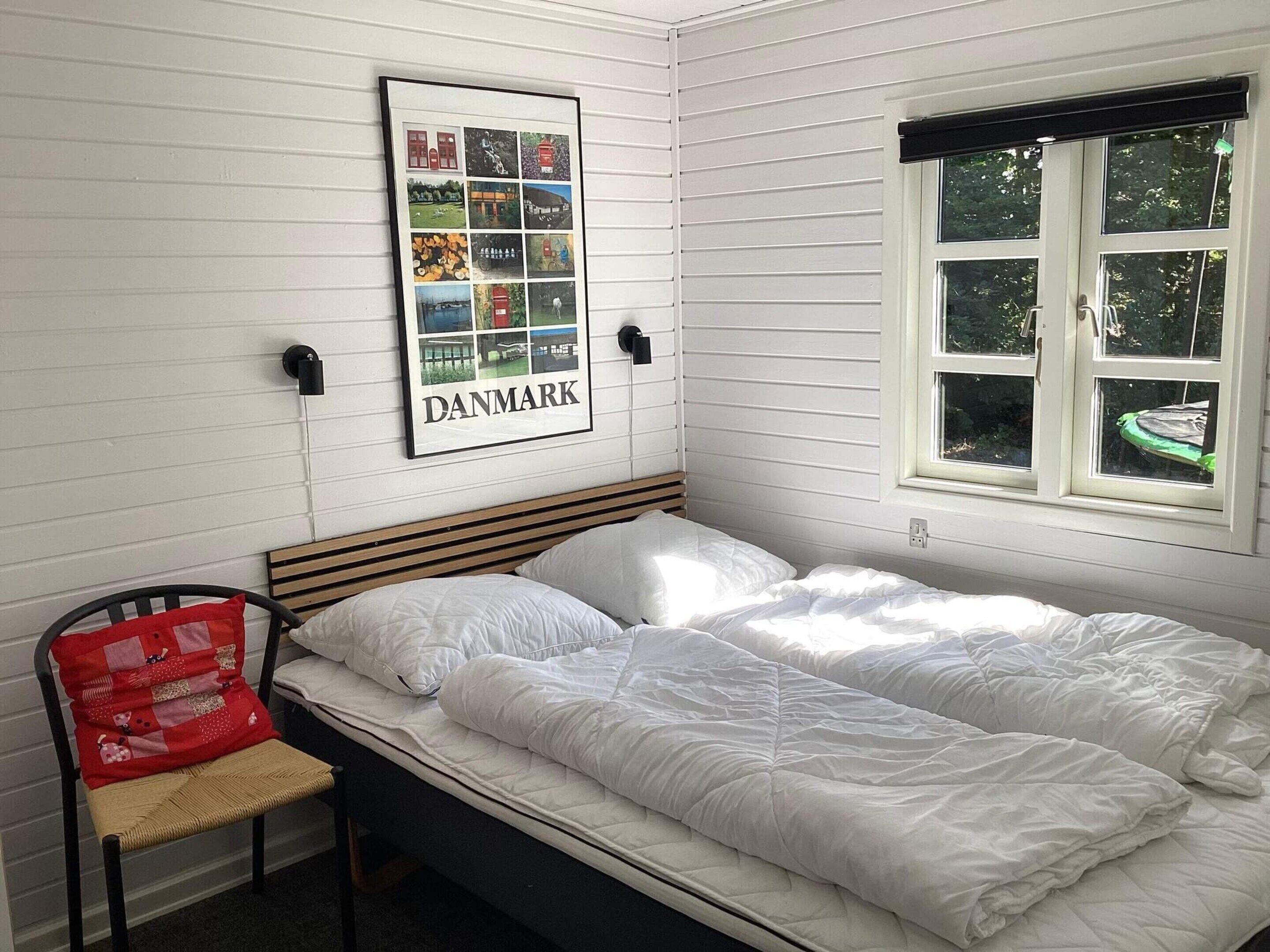 A single-bed room with a framed picture of Denmark, a window with a view of greenery, and a chair with a red cushion.