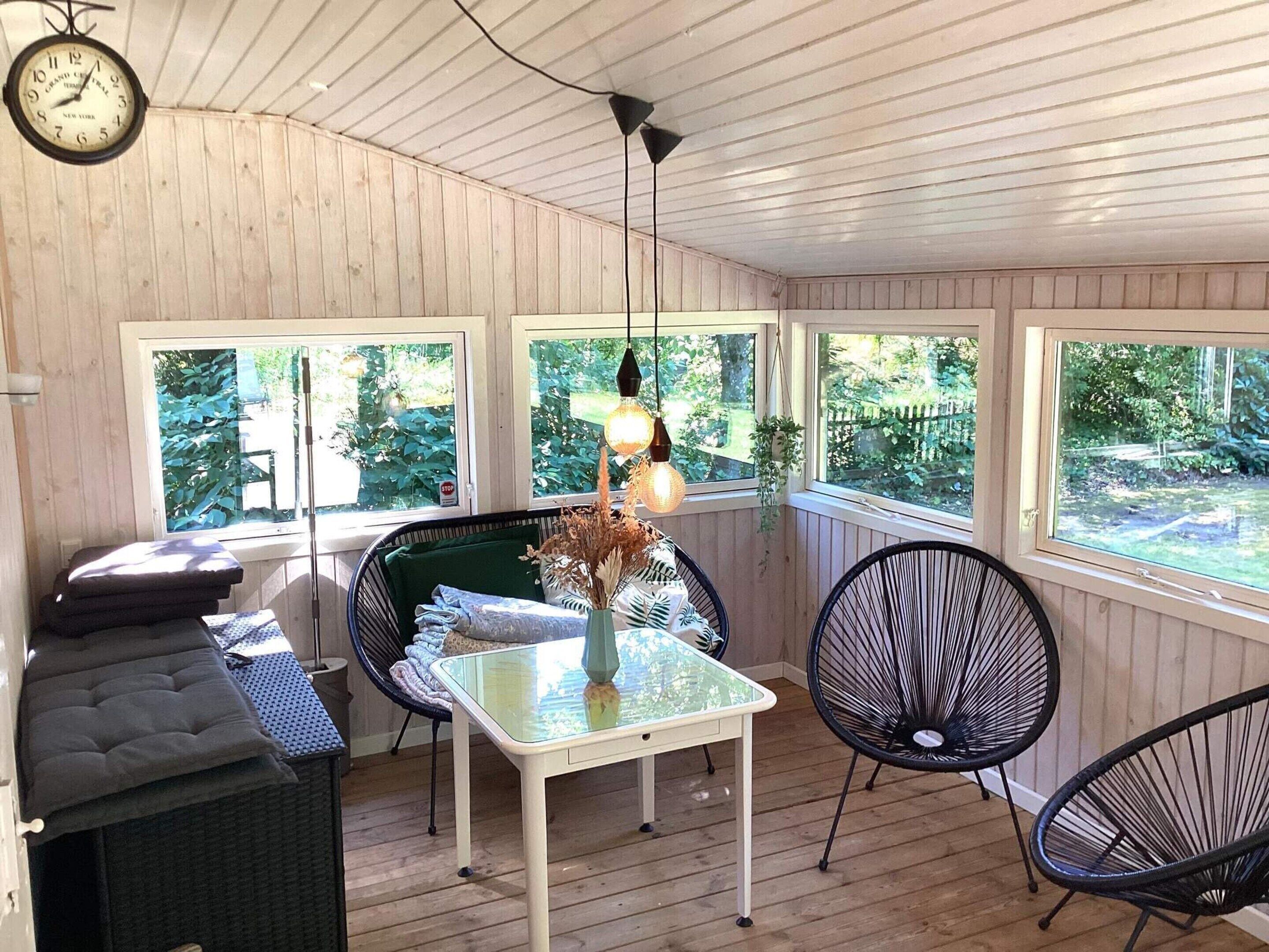A room with a wooden ceiling, a table with chairs, a clock, and windows with a view of greenery.