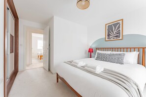 Ein Hotelzimmer mit einem Bett, einem Nachttisch mit Lampe, einem gerahmten Kunstwerk an der Wand und Blick in das angrenzende Zimmer.
