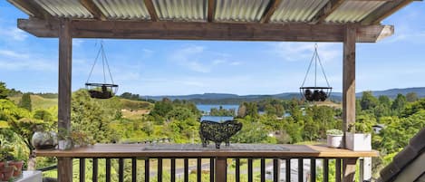 A wooden deck with a view of a lake and hills, a hanging planter, and a wicker sofa.