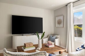 A modern living room with a flat-screen TV mounted on a white wall, a wooden coffee table with books and decorative items, a white sofa with a patterned cushion, and a window with curtains. - 3BR Old Village Retreat - Extended Stay w/ Pool (Mount Pleasant)