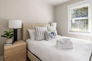 A neatly made bed with pillows, a bedside lamp, a plant, and a window with blinds. - 3BR Old Village Retreat - Extended Stay w/ Pool (Mount Pleasant)