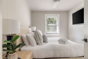 Een slaapkamer met een bed, kussens, een bedlampje, een raam met zonwering, een plant en een aan de muur bevestigde televisie.