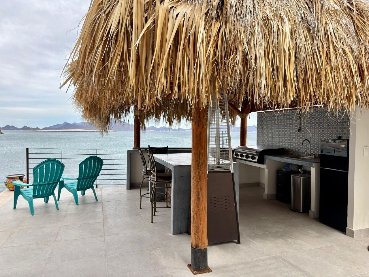 An outdoor kitchen area with a grill, bar stools, and a view of the sea.