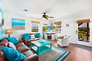 A living room with a brown leather sofa, teal cushions, a teal coffee table, a ceiling fan, a TV on a white cabinet, and a view of the outdoors through a glass door. - Down by the Bay an Oceanside hot tub, and pet-friendly (Kill Devil Hills)