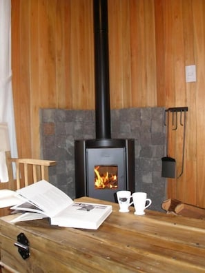 A room with a wood-burning stove, a wooden table with a book and two cups, and a stone fireplace.