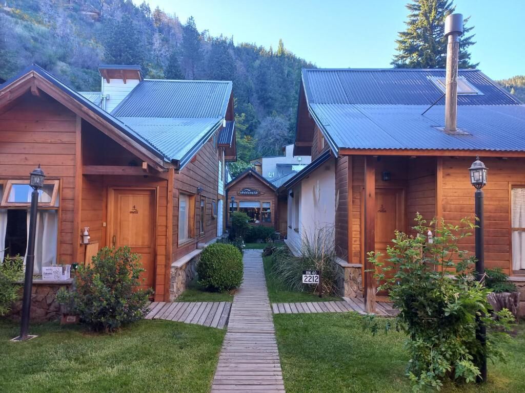 Une rangée de cabanes en bois, disposées de part et d’autre d’un chemin central, entourées de verdure et avec une chaîne de montagnes en arrière-plan.