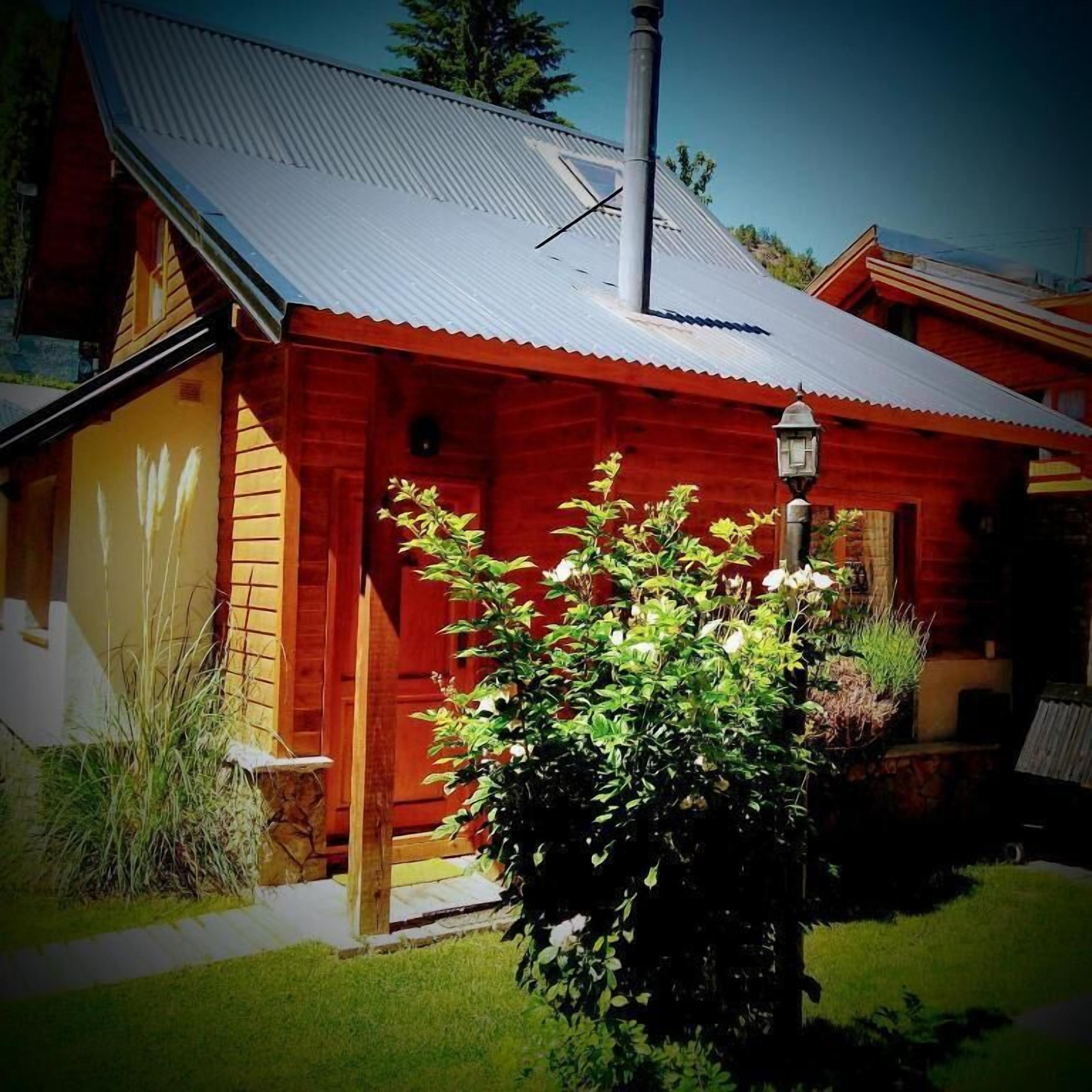 Uma casa de madeira vermelha com telhado de metal, chaminé e um jardim com um arbusto em primeiro plano.