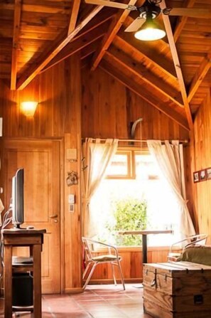 A wooden cabin interior with a bed, a table, and a window with curtains.