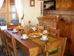 Une table à manger en bois, avec des aliments pour le petit-déjeuner, un vase de fleurs et une vue sur une cuisine dotée de meubles en bois et d’un mur en pierre.