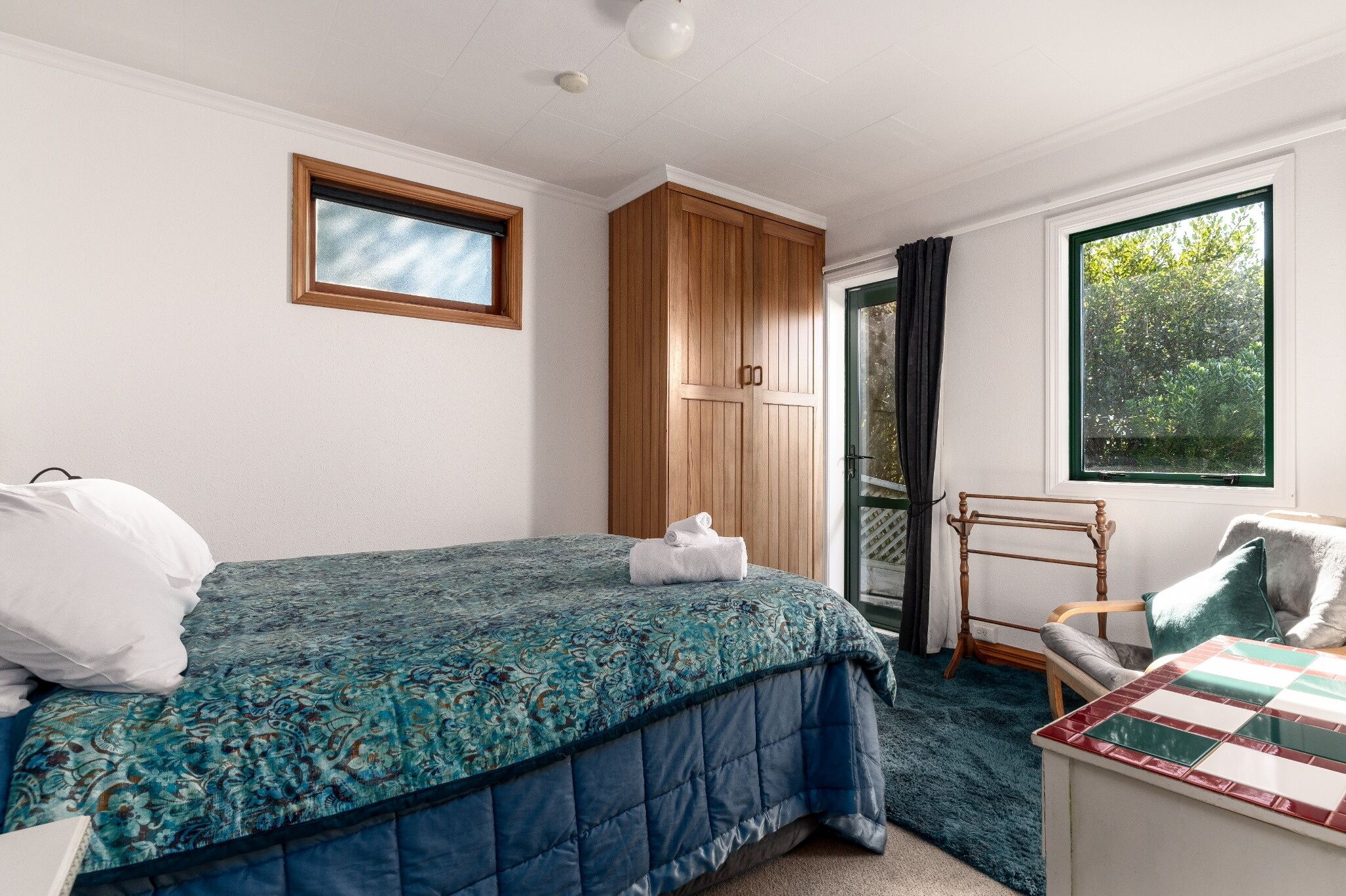 A bedroom with a bed, a wooden wardrobe, a window with a view, and a small table with a chair.