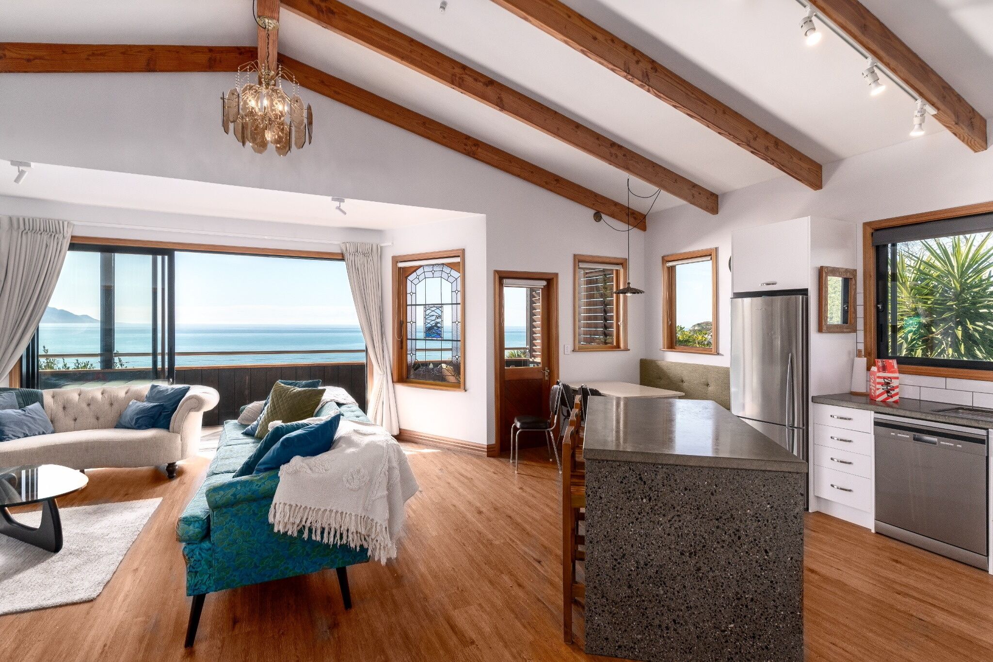 A spacious living area with a view of the sea, a cozy seating area, a kitchen with a modern island, and wooden beams on the ceiling.