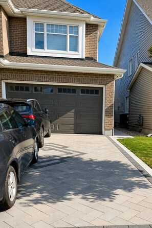 A residential driveway with two cars parked, a garage door, and a house with a brick facade and a window above the garage.