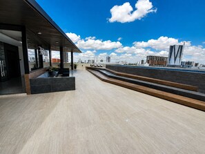 Une terrasse moderne sur le toit, offrant une vue sur les gratte-ciel de la ville, avec un espace salon couvert et un escalier menant à l’étage supérieur.