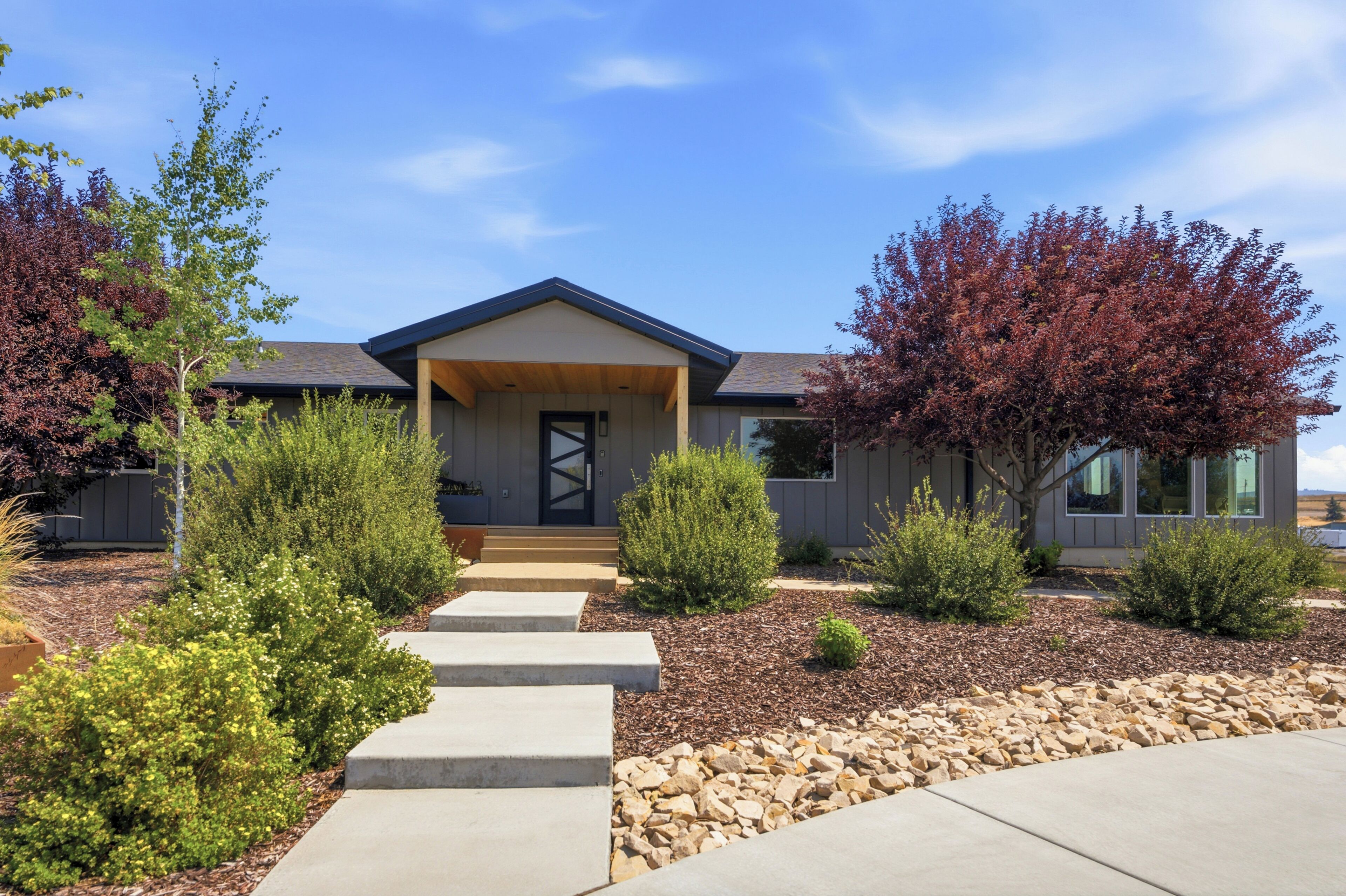 A modern two-story house with a front porch, a stone pathway leading to the door, and a landscaped garden with various shrubs and trees.
