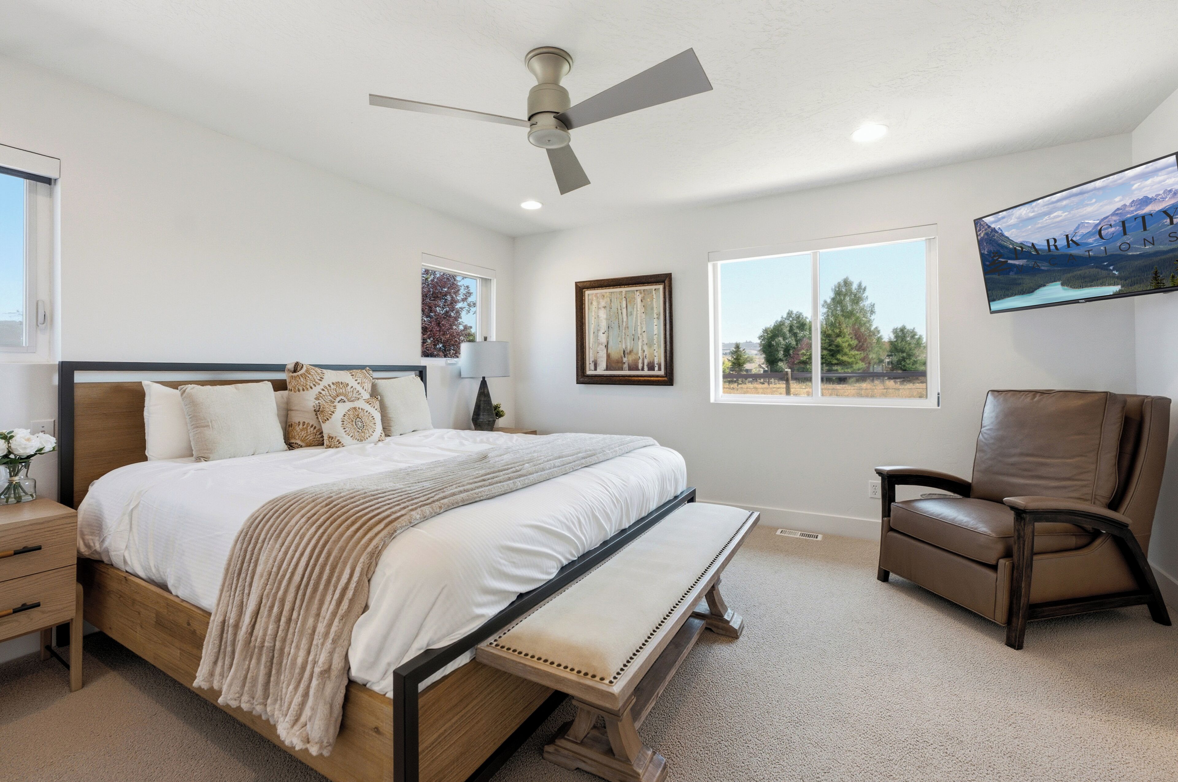 A bedroom with a bed, a bench, a chair, a ceiling fan, and a painting on the wall.