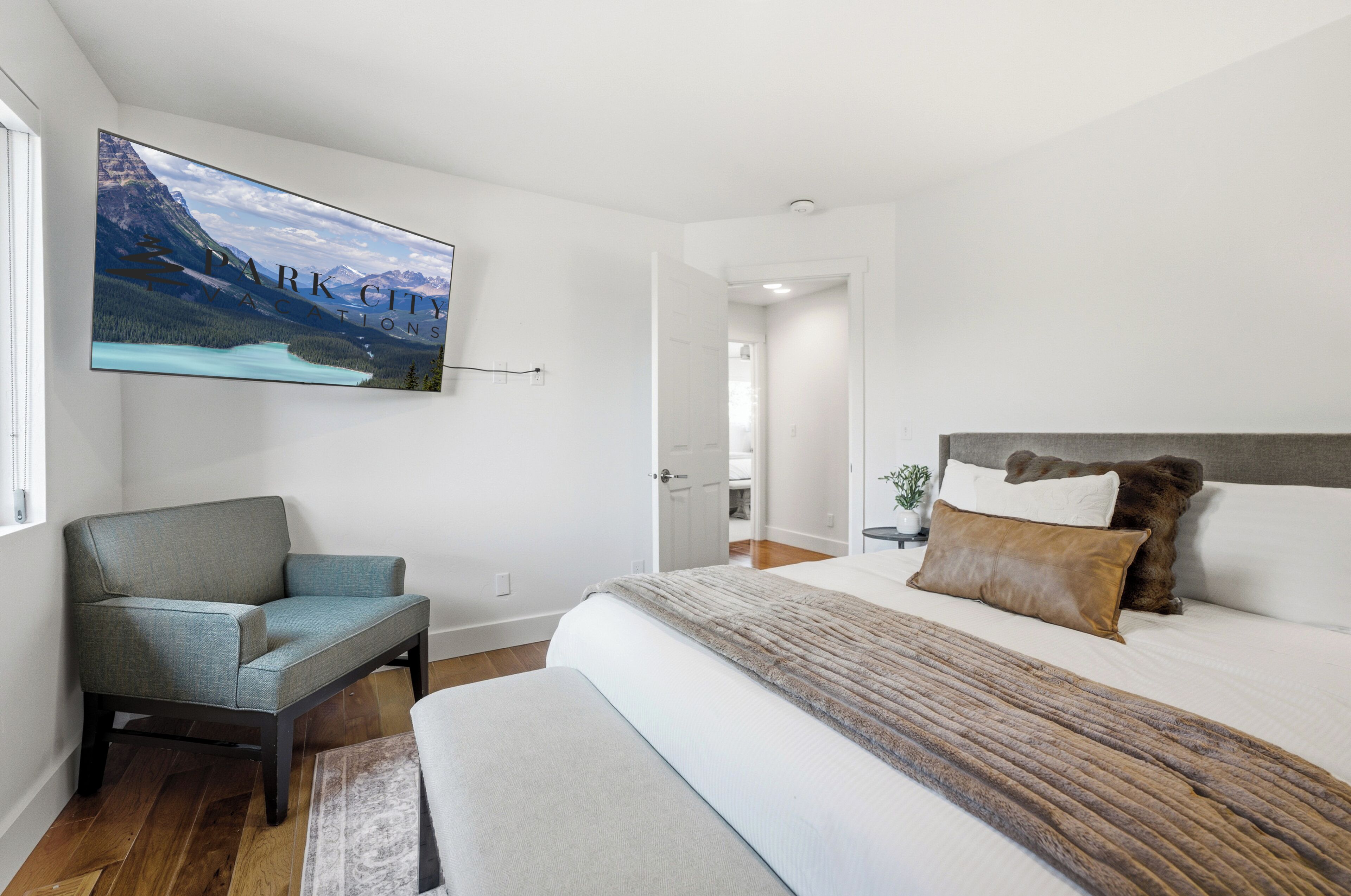 A modern bedroom with a large bed, a chair, a TV mounted on the wall, and a view of a mountain landscape.