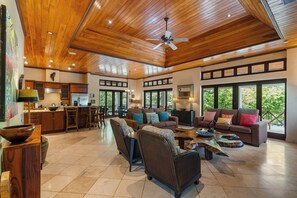 A spacious living area with a high ceiling, wooden flooring, and a large window offering a view of greenery. - 6 Bdr Penthouse with Plunge Pool and Beach Club Access! (Tamarindo)