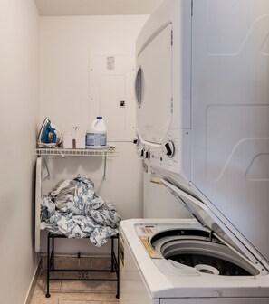 A compact laundry room with a front-loading washing machine, a drying rack, and a storage shelf. - Sunshine on Crestdale - 3/2 Pool home Sleeps 8 (Palm Beach Gardens)