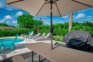 Ein Außenpoolbereich mit Liegestühlen, einem überdachten Grill und einem Tisch unter einem großen Sonnenschirm.