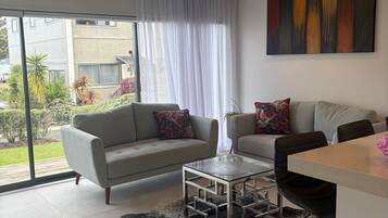 Una sala de estar moderna con un sofá, una mesa de centro de vidrio y un comedor con un taburete de bar.