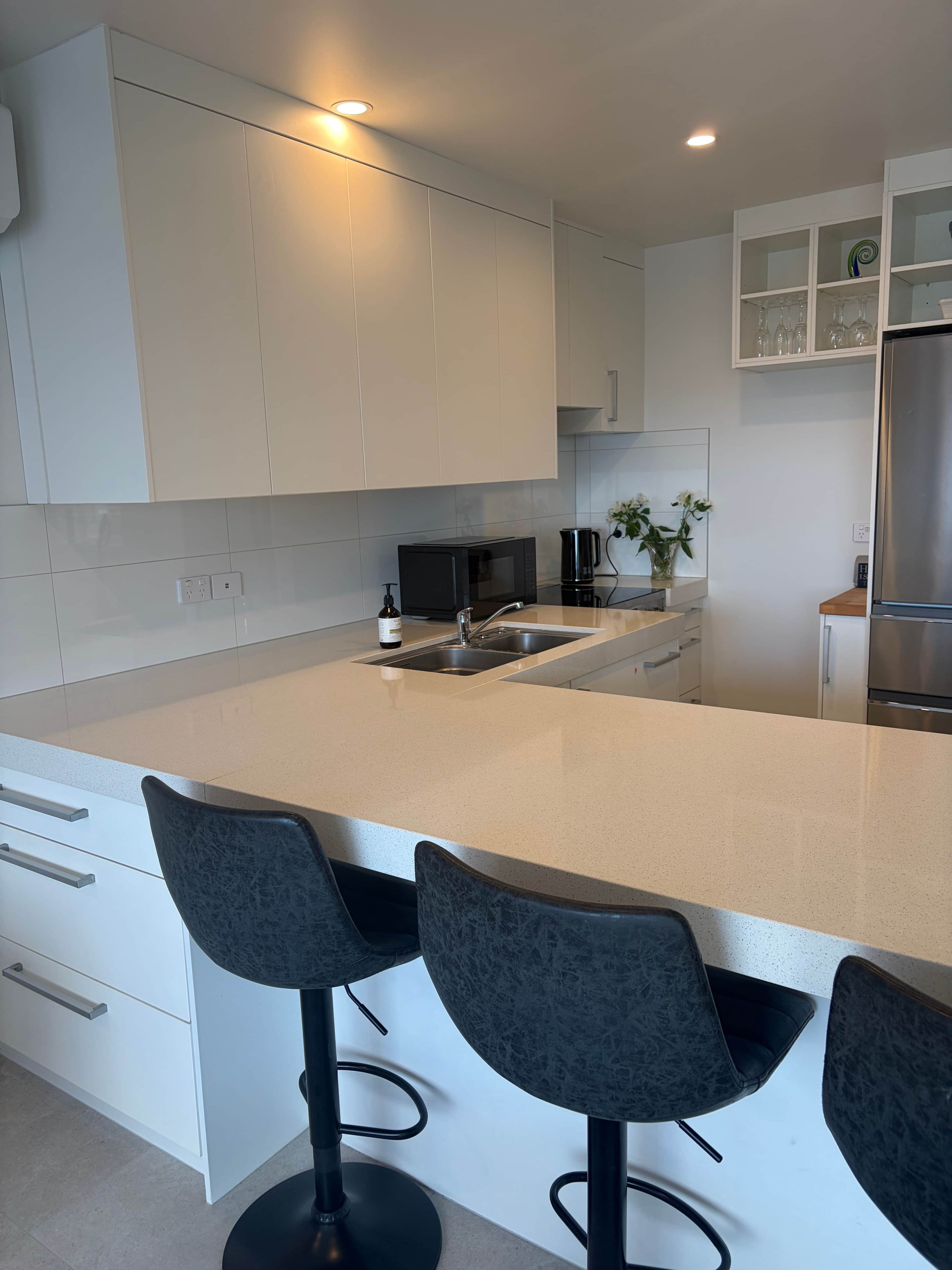 A modern kitchen with white cabinetry, a stainless steel refrigerator, a microwave, and a sink. There are two black bar stools with a round table in front.