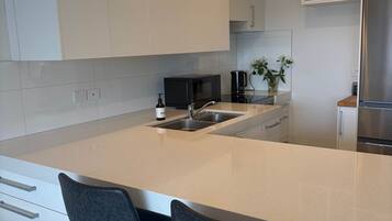A modern kitchen with white cabinetry, a stainless steel refrigerator, a microwave, and a sink. There are two black bar stools with a round table in front.