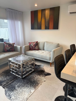 A modern living room with a sofa, a glass coffee table, and a dining area with chairs. - Sandy Soul Beach Apartment (Whitianga)