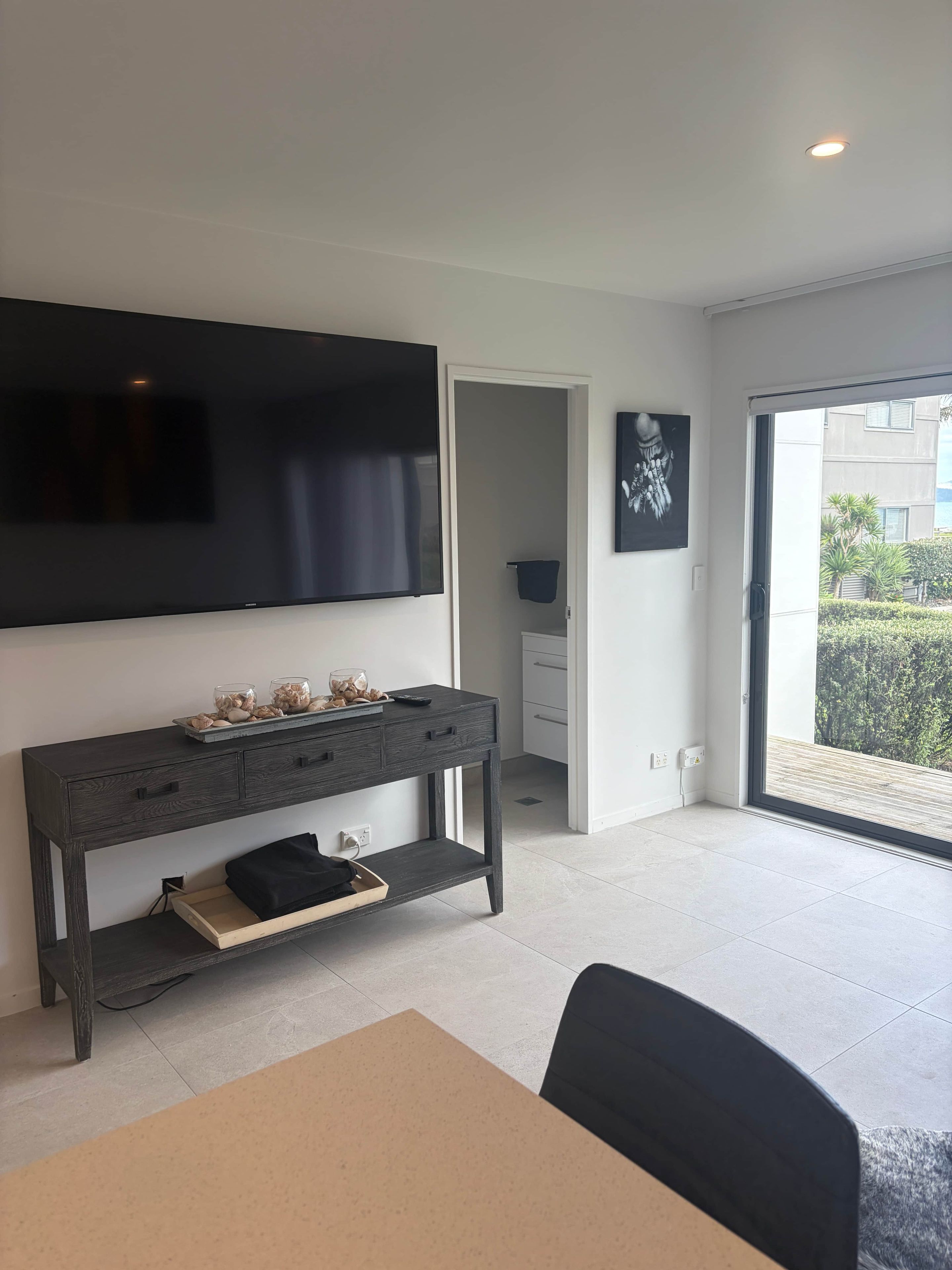 Una sala moderna con un televisor de pantalla plana montado en la pared, una consola de madera oscura con objetos decorativos, una zona de comedor con mesa y sillas, y un gran ventanal corredizo que da a un balcón exterior.