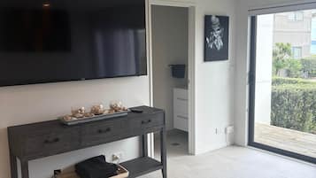 Una sala moderna con un televisor de pantalla plana montado en la pared, una consola de madera oscura con objetos decorativos, una zona de comedor con mesa y sillas, y un gran ventanal corredizo que da a un balcón exterior.