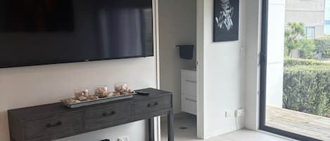 Un salon moderne avec un téléviseur à écran plat fixé au mur, une console en bois foncé agrémentée d’objets décoratifs, un coin repas avec une table et des chaises, et une grande porte-fenêtre coulissante donnant sur un balcon extérieur.
