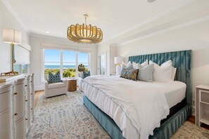 A bedroom with a large bed, a view of the ocean, a chandelier, and a dresser. - Oceanfront 5BR home with private pool, hot tub, boardwalk & beach access. (Isle Of Palms)