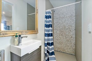 A bathroom with a white sink, a mirror with an ornate frame, a shower with a pebble-textured wall, and a striped shower curtain. - Sandy Soul Beach Studio (Whitianga)